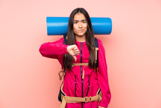 Young mountaineer Indian girl with a big backpack isolated on pink background showing thumb down sign