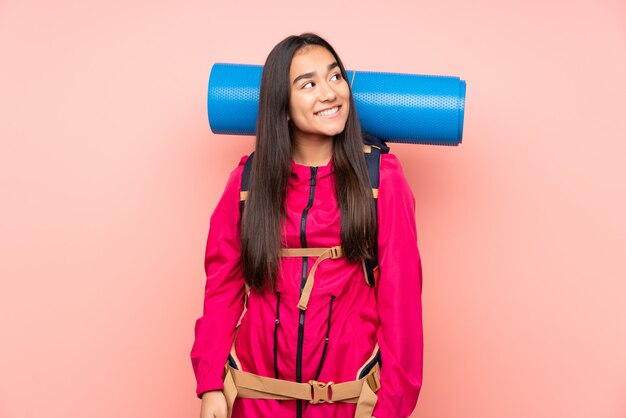 Young mountaineer Indian girl with a big backpack on pink wall laughing and looking up