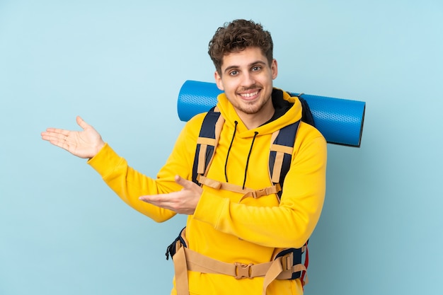 Young mountaineer man with a big backpack on blue wall extending hands to the side