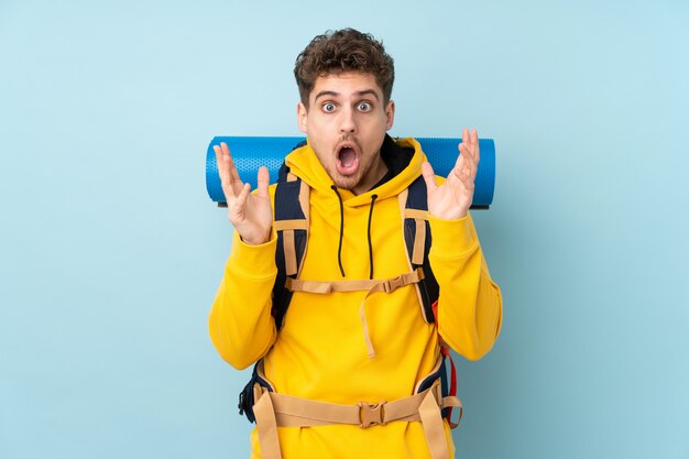 Young mountaineer man with a big backpack on blue wall with surprised facial expression