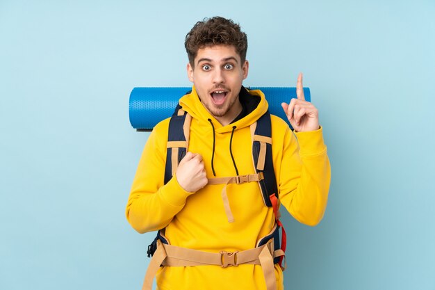 Young mountaineer man with a big backpack on blue wall with surprised facial expression