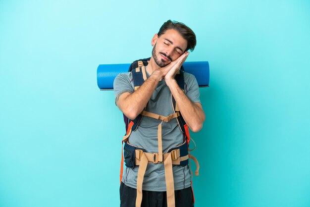 Young mountaineer man with a big backpack isolated on blue background making sleep gesture in dorable expression