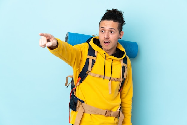 Young mountaineer man with a big backpack over isolated blue background pointing away