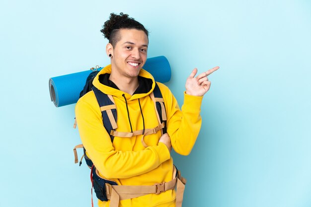 Young mountaineer man with a big backpack over isolated blue background pointing finger to the side