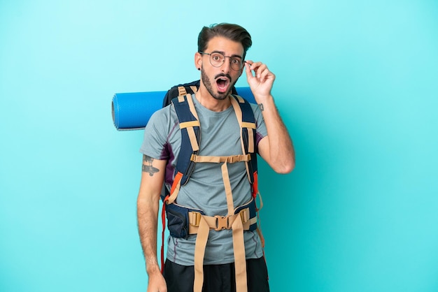 Молодой альпинист с большим рюкзаком изолирован на синем фоне в очках и удивлен