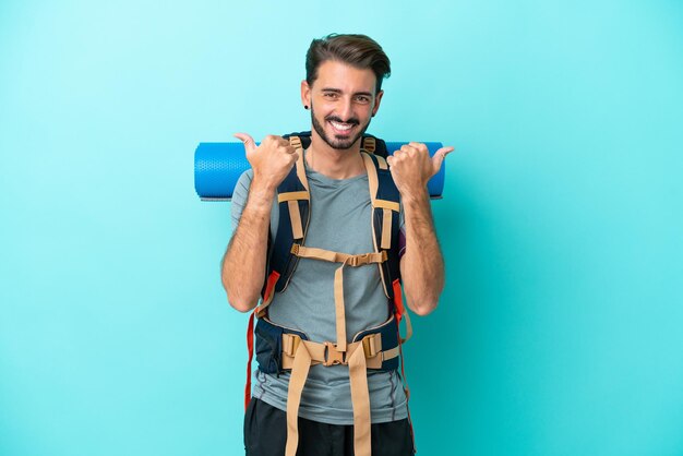 Young mountaineer man with a big backpack isolated on blue background with thumbs up gesture and smiling