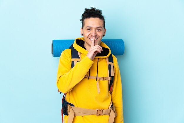 Young mountaineer man with a big backpack over isolated blue wall doing silence gesture