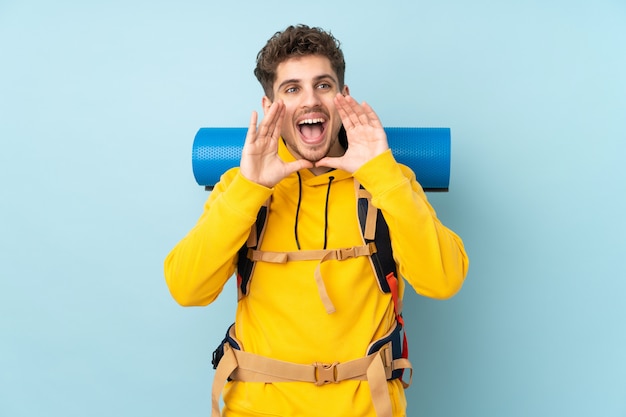 Young mountaineer man with a big backpack isolated on blue  wall shouting with mouth wide open