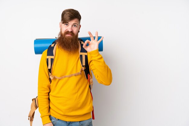 Young mountaineer man with a big backpack over isolated white wall showing an ok sign with fingers