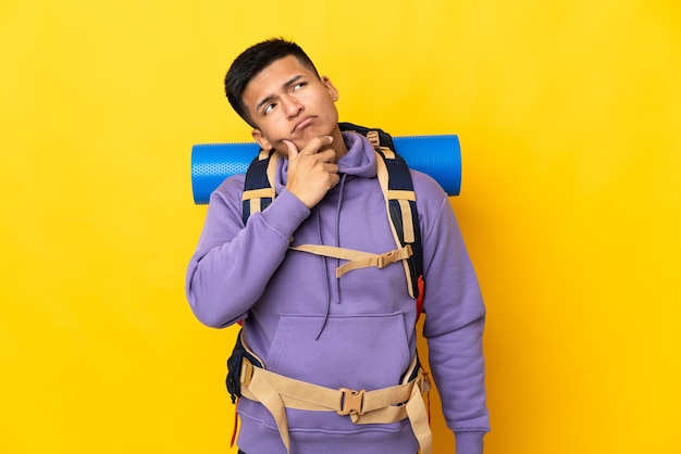 Young mountaineer man with a big backpack isolated on yellow background having doubts