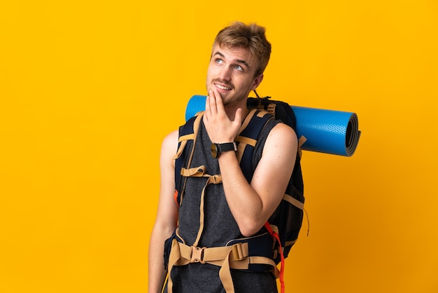 Young mountaineer man with a big backpack isolated on yellow background looking up while smiling