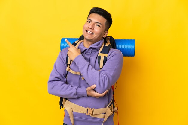 Young mountaineer man with a big backpack isolated on yellow background pointing to the side to present a product