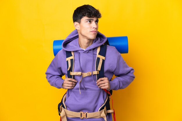 Young mountaineer man with a big backpack on isolated yellow and looking up