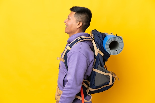 Young mountaineer man with a big backpack isolated on yellow wall laughing in lateral position