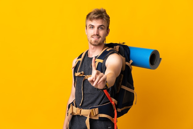 Молодой альпинист с большим рюкзаком на желтой стене показывает и поднимает палец