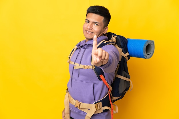 Young mountaineer man with a big backpack isolated on yellow wall showing and lifting a finger