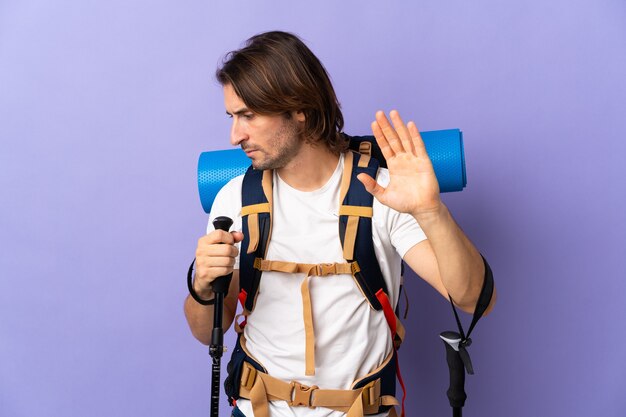 Young mountaineer man with a big backpack making stop gesture and disappointed