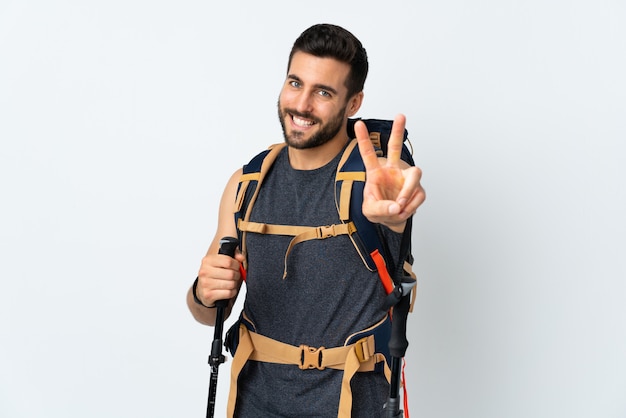 Uomo giovane alpinista con un grande zaino e bastoncini da trekking isolato su bianco sorridente e mostrando il segno della vittoria