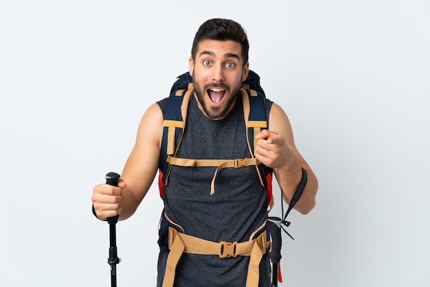 Young mountaineer man with a big backpack and trekking poles isolated on white wall points finger at you