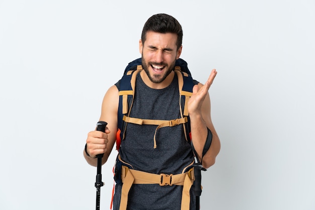 Young mountaineer man with a big backpack and trekking poles on white wall laughing