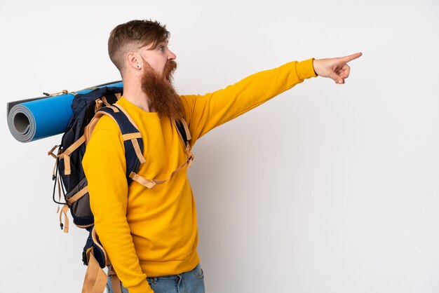 Young mountaineer man with a big backpack over white wall pointing finger to the side