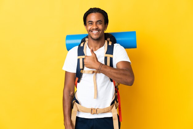 Young mountaineer man with braids with a big backpack isolated on yellow wall giving a thumbs up gesture