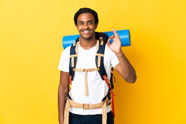 Young mountaineer man with braids with a big backpack isolated on yellow wall showing ok sign with fingers