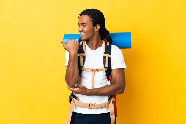 Young mountaineer man with braids with a big backpack on yellow pointing to the side to present a product