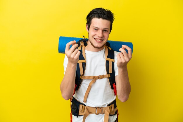 Young mountaineer Russian man with a big backpack isolated on yellow background making money gesture