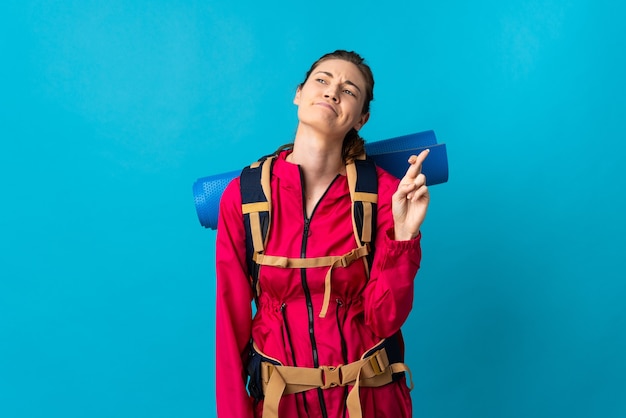 Young mountaineer woman over isolated blue background with fingers crossing and wishing the best