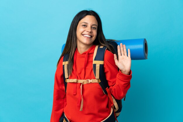 Young mountaineer woman with backpack saluting with hand with happy expression