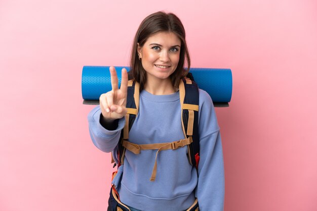 Giovane donna alpinista con un grande zaino isolato su sfondo rosa che sorride e mostra il segno della vittoria