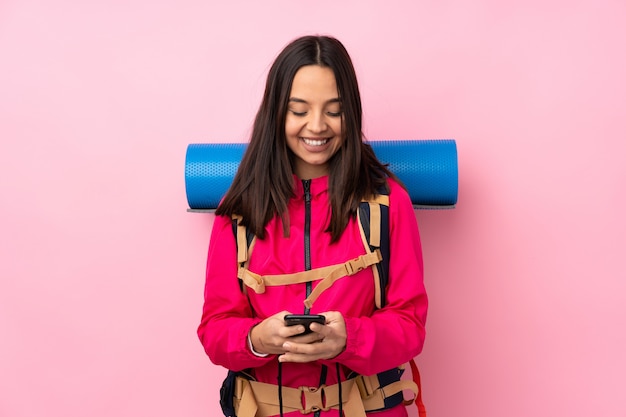 Young mountaineer woman with a big backpack over isolated pink wall sending a message with the mobile