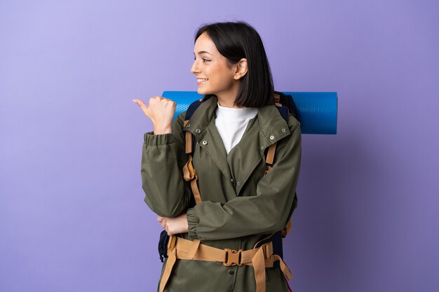 Young mountaineer woman with a big backpack over isolated pointing to the side to present a product