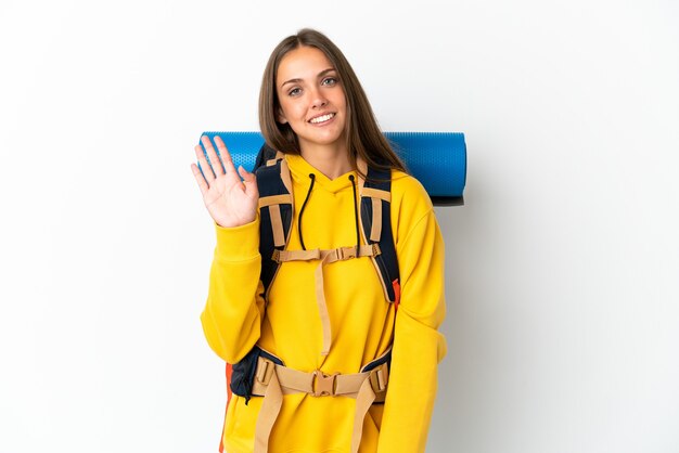 Young mountaineer woman with a big backpack over isolated white background saluting with hand with happy expression