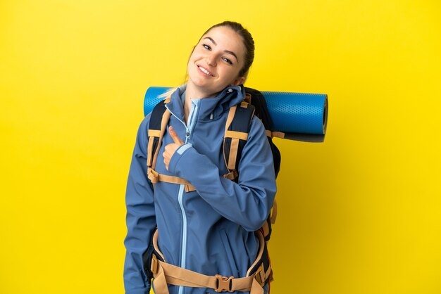 Young mountaineer woman with a big backpack over isolated yellow background giving a thumbs up gesture