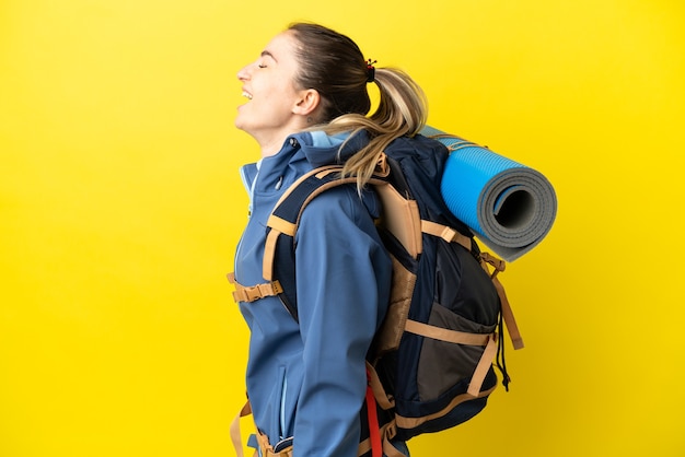 Молодая альпинистка с большим рюкзаком на изолированном желтом фоне смеется в боковом положении