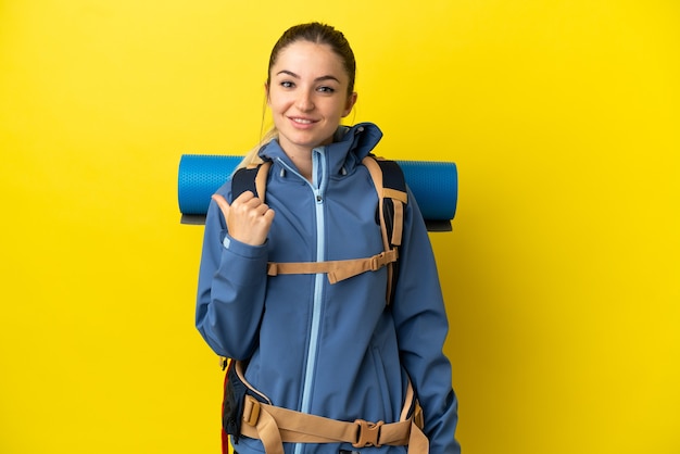 Young mountaineer woman with a big backpack over isolated yellow background pointing to the side to present a product