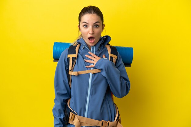 Young mountaineer woman with a big backpack over isolated yellow background surprised and shocked while looking right