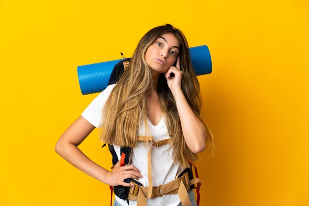 Young mountaineer woman with a big backpack isolated on yellow wall thinking an idea