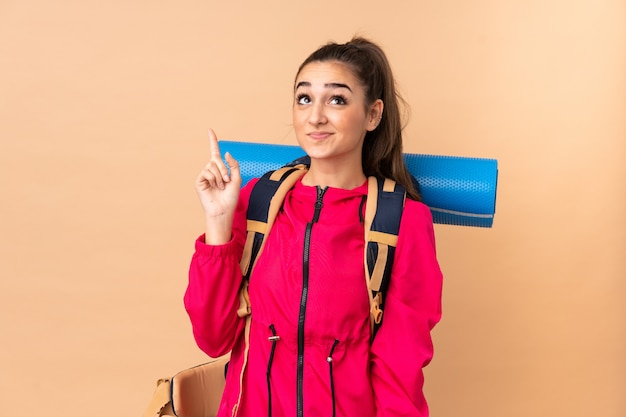 Young mountaineer woman with a big backpacker isolated on beige wall pointing with the index finger a great idea