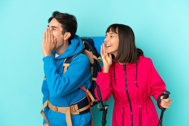 Young mountaineers couple isolated on blue background shouting with mouth wide open to the lateral