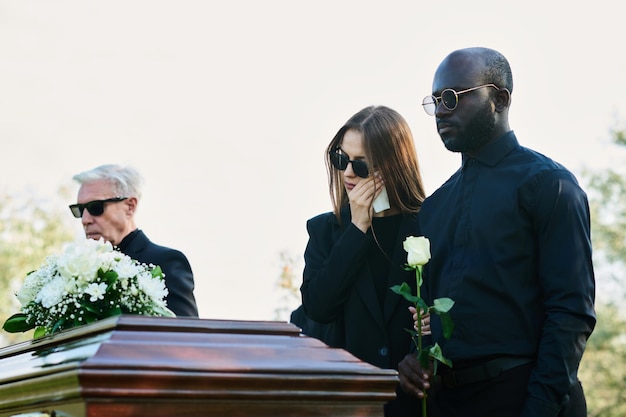 Young mourning man with white rose standing by his wife wiping tears