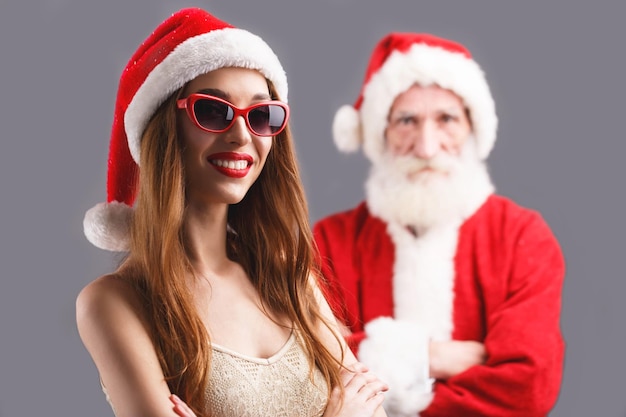 Молодая миссис Клаус в шляпе Санта-Клауса и солнцезащитных очках стоит и улыбается Санта-Клаусу