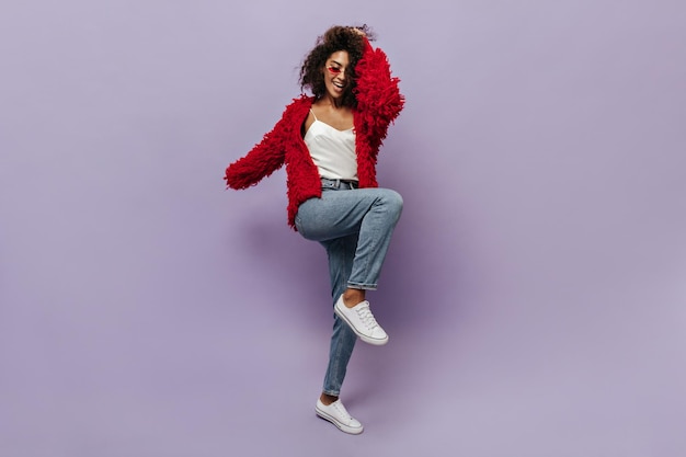 Young mulatto girl with wavy hairstyle in bright fluffy cool clothes and white stylish sneakers posing with cute smile on lilac backdrop