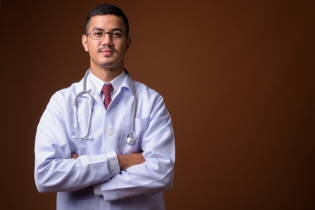 Young multi-ethnic Asian man doctor against brown wall