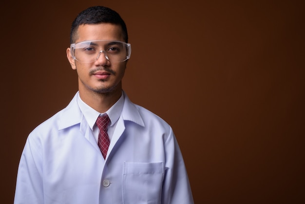 Young multi-ethnic Asian man doctor against brown wall