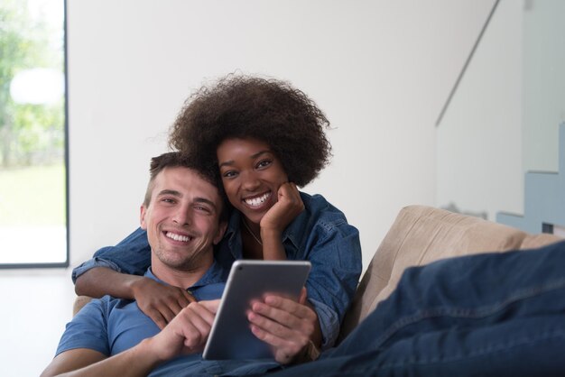 Young multiethnic couple relaxing at luxurious home with tablet computers reading in the living room on the sofa couch.