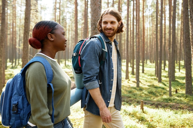 Молодая многонациональная пара с рюкзаками гуляет и разговаривает вместе в лесу