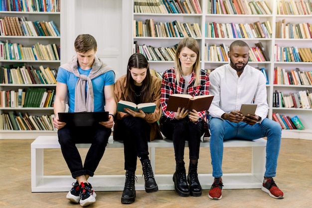 Young multiethnic smart students, girls and boys, reading in library from traditional textbooks and ebook and laptop